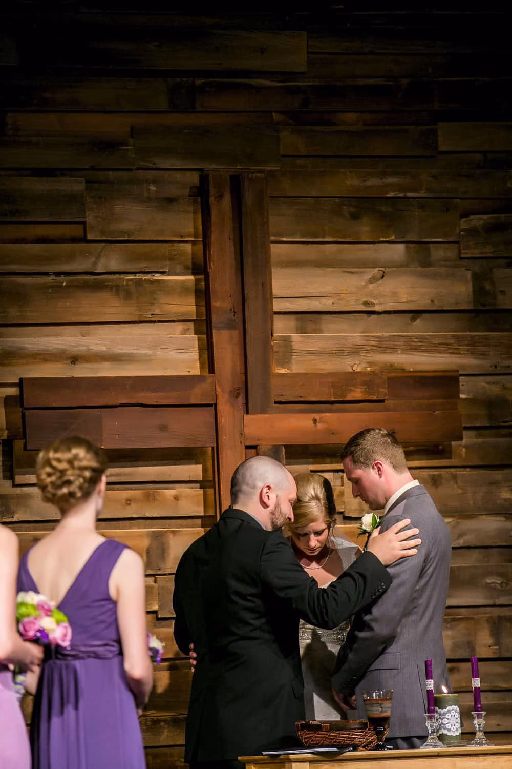 A couple stands at the altar holding hands as a man in a suit leads a prayer, while two bridesmaids in purple dresses stand nearby before a large wooden cross—captured beautifully by Madison Wedding Photography.