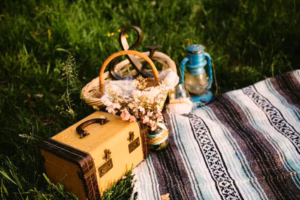A woven blanket on grass with a picnic basket, blue lantern, bouquet of flowers in a jar, and a small vintage suitcase—perfect inspiration for any Eau Claire wedding photographer capturing dreamy outdoor moments.