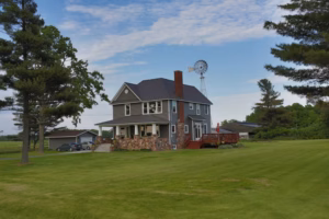 A gray two-story house with a stone porch and red deck sits on a large grassy lawn—an ideal backdrop for an Eau Claire Wedding Photographer—with trees, a windmill, and a detached garage under a partly cloudy sky.