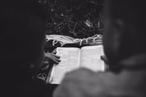 Two people sit outdoors closely together, reading an open book placed on their laps, with one hand resting on the page. Captured in black and white by an Eau Claire wedding photographer.