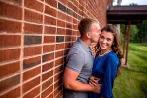 A man in a gray shirt kisses a woman in a blue dress on the forehead as they stand against a brick wall, with greenery and a building in the background—captured beautifully by an Eau Claire Wedding Photographer.