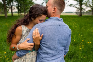A woman embraces a man outdoors in a grassy field, resting her head on his shoulder while he kisses her forehead—captured beautifully by an Eau Claire wedding photographer.