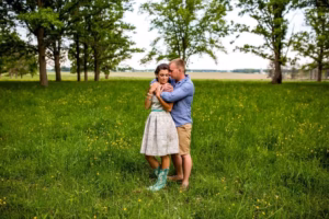 A man and woman stand close together in a green field with trees, the woman in a dress and cowboy boots, the man in a blue shirt and shorts—captured tenderly by an Eau Claire Wedding Photographer as they both look down.