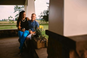 A woman and a man sit together on a porch, holding drinks and smiling, with a potted plant and green fields in the background—captured beautifully by an Eau Claire wedding photographer.