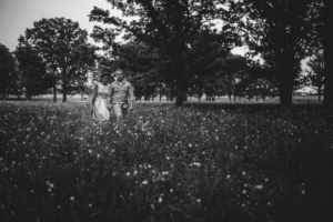 A couple walks through a field of wildflowers with tall trees in the background, captured in black and white by an Eau Claire Wedding Photographer.