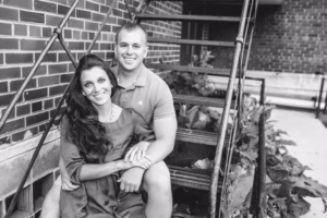 A couple sits together on outdoor metal stairs by a brick building, smiling at the camera. Captured in black and white by an Eau Claire Wedding Photographer, this moment radiates timeless love and joy.