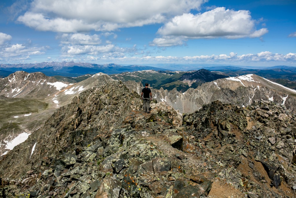 A man conquering Quandary, a 14er, while standing on top of a mountain with other peaks in the background.