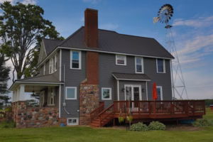 Two-story house with gray siding, stone foundation, large chimney, covered porch, wooden deck with red umbrella—a perfect photo spot for any Eau Claire Wedding Photographer—with a charming windmill in the background.