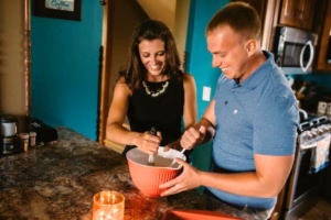 A woman and a man stand in a kitchen, smiling while mixing ingredients in a large red bowl on the countertop next to a lit candle, perfectly capturing the candid joy an Eau Claire Wedding Photographer might seek.