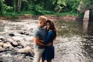 A couple stands closely together on rocks by a river in a forested area, with the water and trees visible in the background, beautifully captured by an Eau Claire wedding photographer.