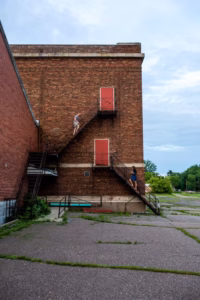 Two people walk on opposite flights of an exterior metal staircase attached to a brick building with red doors, as if captured through the artistic eye of an Eau Claire wedding photographer, surrounded by mostly empty, cracked pavement.