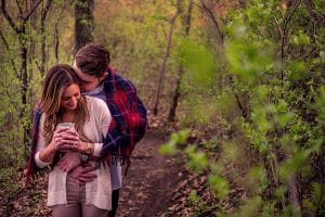An adventurous couple captures engagement photos while embracing in the woods, cell phone in hand.