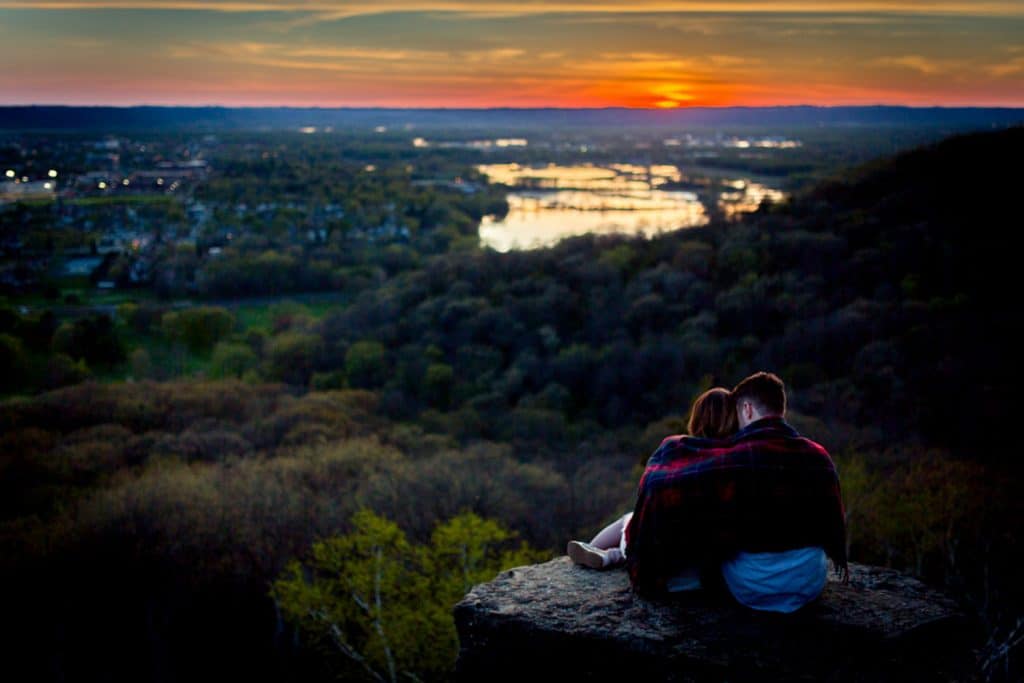 An adventurous couple captures their engagement photos on a rock, admiring the cityscape during a picturesque sunset.