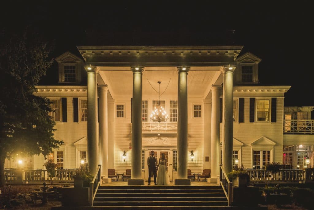 A bride and groom captured by a skilled Littleton Wedding Photographer in front of a large mansion at night.