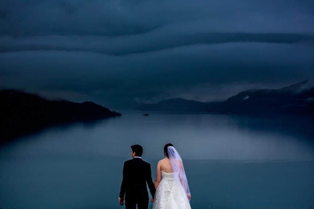 A Jacks Point Queenstown wedding held under a stormy sky with the bride and groom standing in front of a lake.