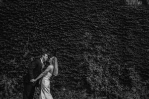 A bride and groom kiss in front of an ivy covered wall.