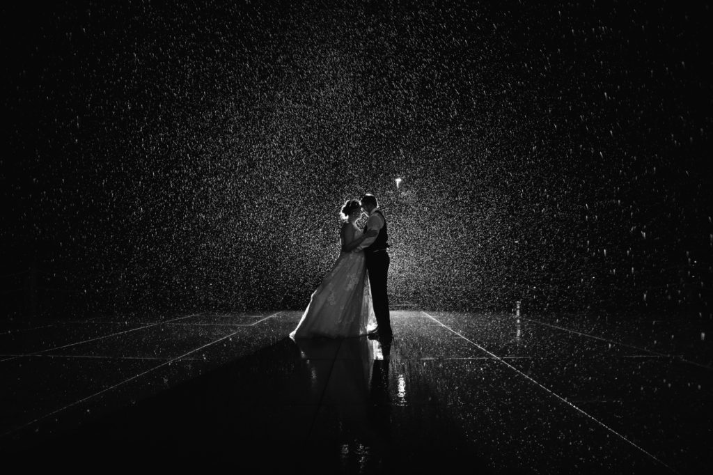 A bride and groom standing in the rain in a dark room.