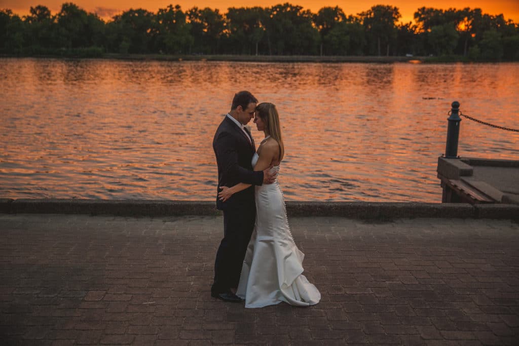 A Charmant Wedding couple embracing in front of a river at sunset.