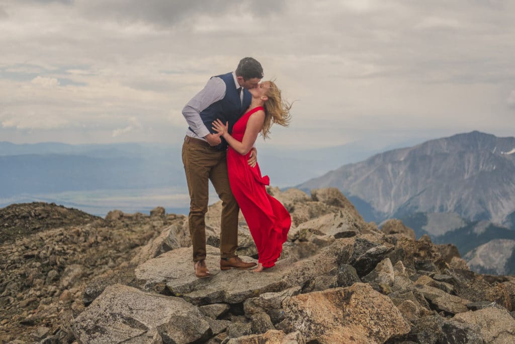 An engaged couple kissing on top of a mountain.