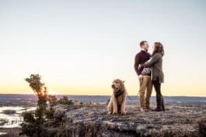 A couple and their dog standing on top of a cliff at sunset.