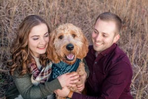 A man and woman pose with their dog in a field.