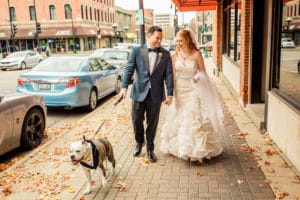 A bride and groom, accompanied by their dog, stroll down the street near 4 Sisters Wine Bar on their wedding day.