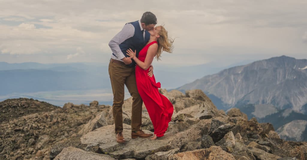 A couple kissing on top of a mountain in a red dress.