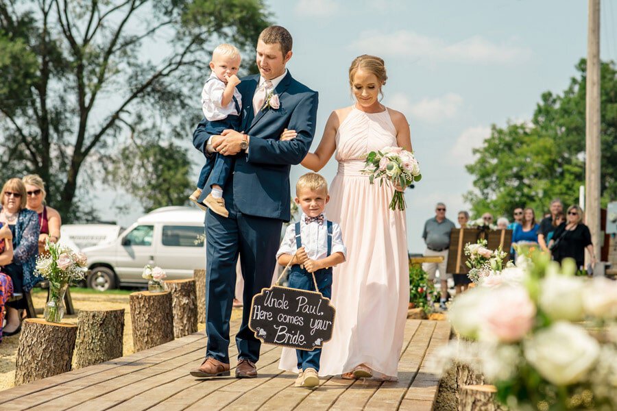 A bride and groom, with their children in tow, elegantly stroll down a charming wooden walkway at their blissful On A Whim Barn Wedding.