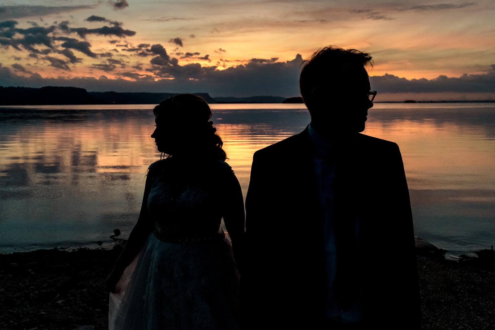 A silhouette of a bride and groom standing by a body of water at sunset.