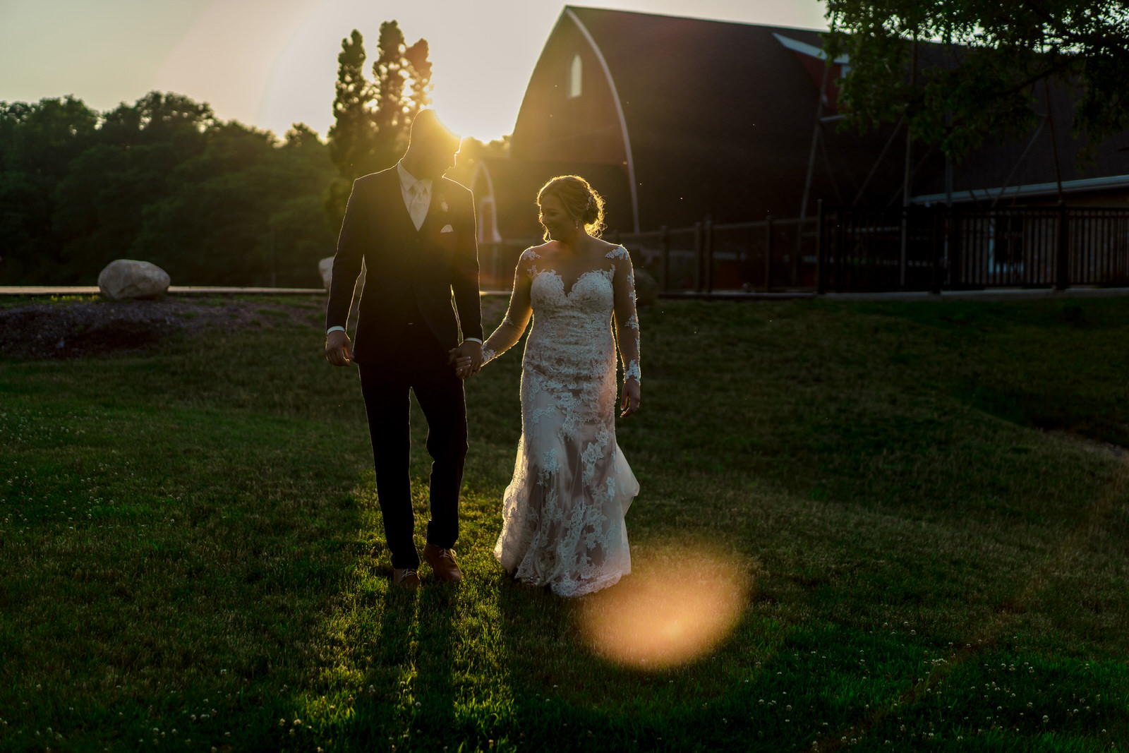 A bride and groom walking in front of a barn at sunset.