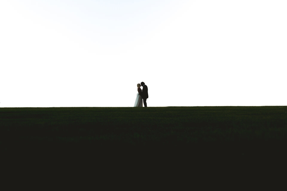 A silhouette of a bride and groom standing on a grassy field.