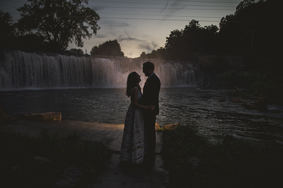 A bride and groom standing in front of a waterfall at sunset.