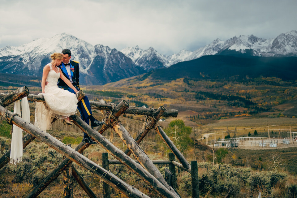 A bride and groom standing on a wooden bridge with mountains in the background.