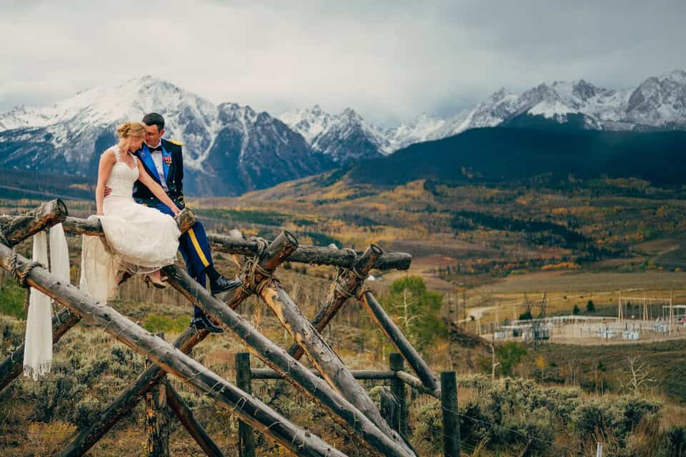 A bride and groom standing on a wooden bridge with mountains in the background.