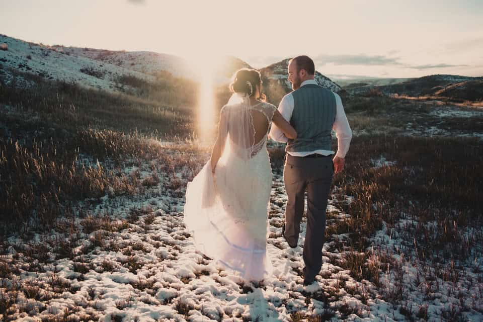 A bride and groom walking through a snowy field at sunset.