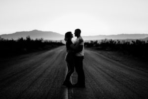 A black and white photo of a couple standing in the middle of a desert road.