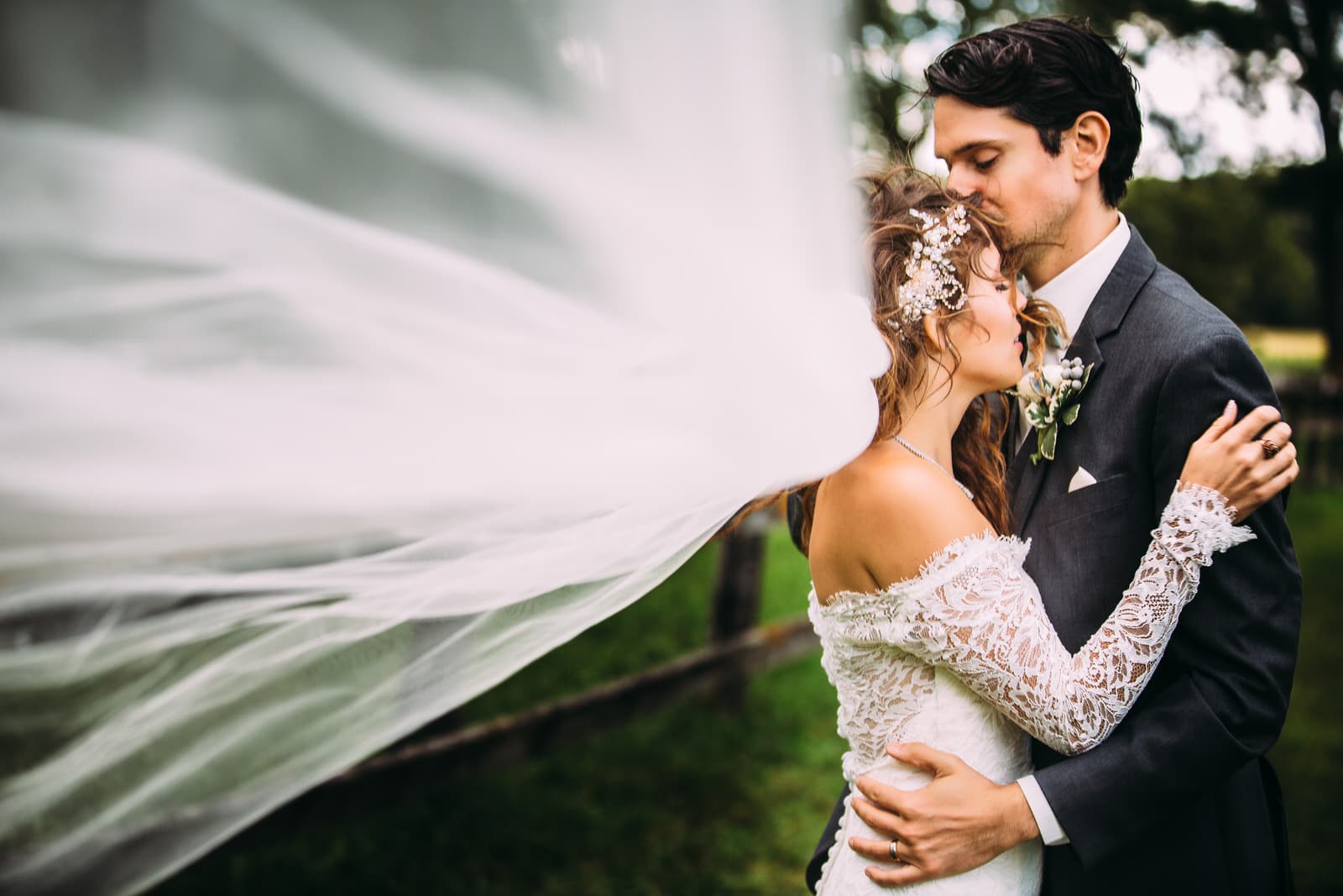 A bride and groom embrace under a veil in a field.