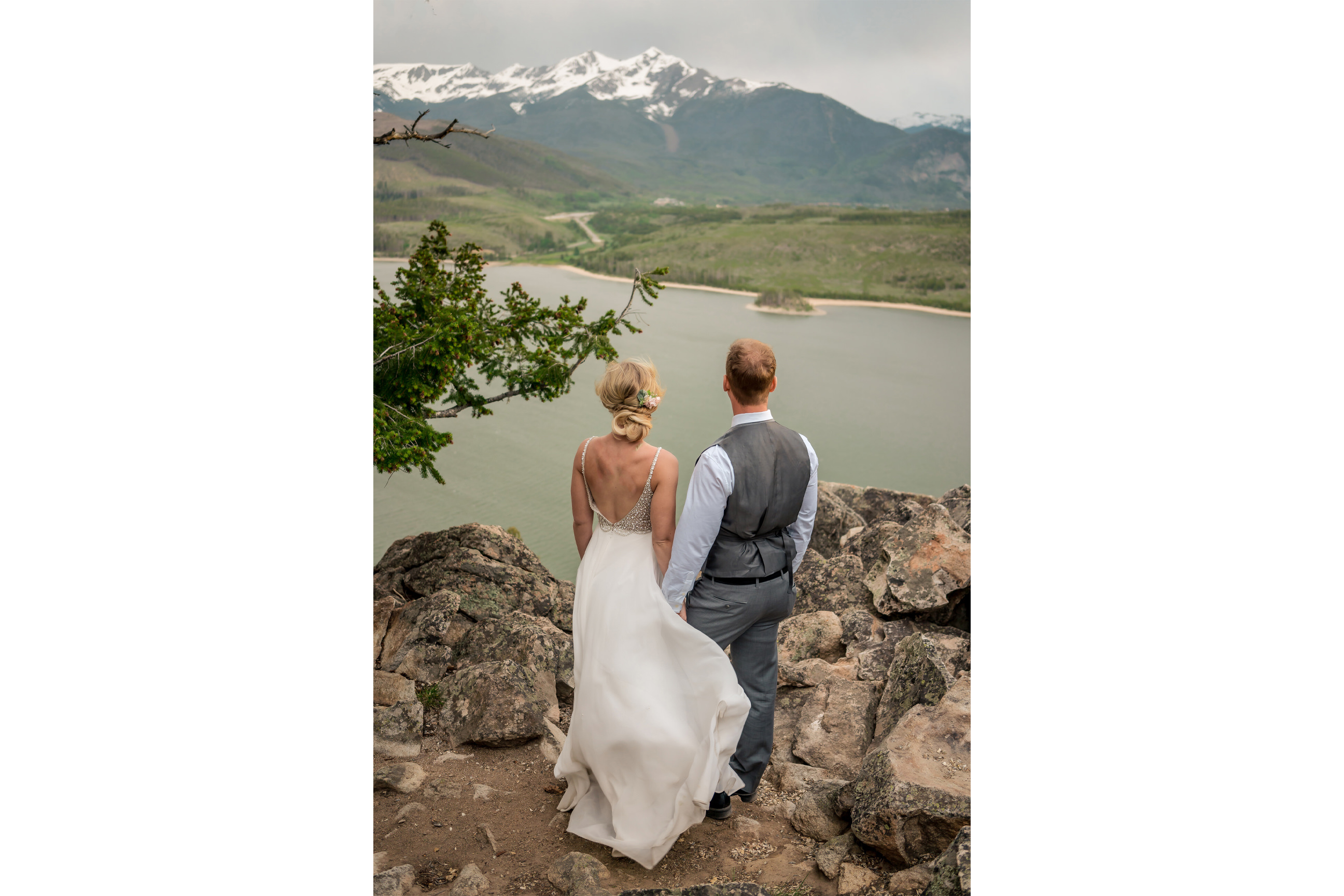 A bride and groom standing on top of a mountain overlooking a lake.