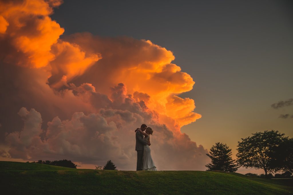 A bride and groom kiss in front of a cloud at sunset.