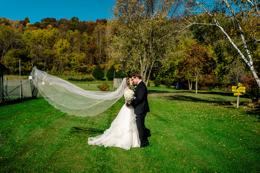 A bride and groom kissing in the grass with their veil blowing in the wind.