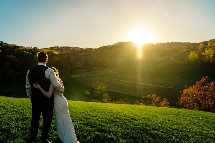 A bride and groom standing in a field with the sun setting behind them.