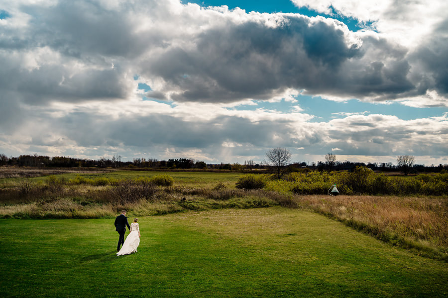 A bride and groom walking in a field under a cloudy sky.
