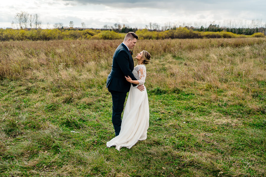 A bride and groom embrace in a field under a cloudy sky.