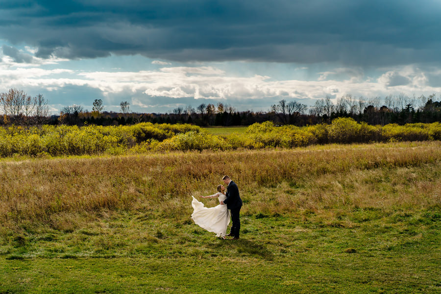 A bride and groom standing in a field under a cloudy sky.