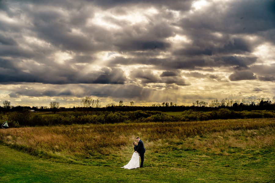 A bride and groom standing in a field under a stormy sky.