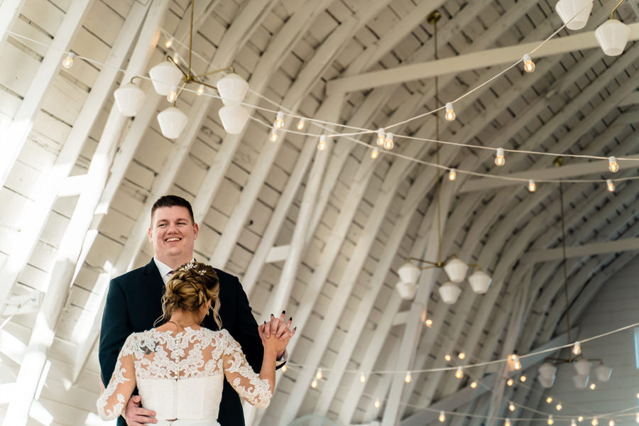 A bride and groom standing in a white barn with string lights.