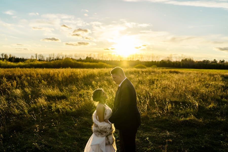 A bride and groom standing in a field at sunset.