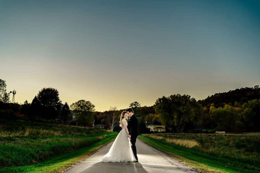 A bride and groom standing on a dirt road at sunset.