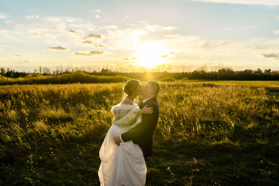 A bride and groom embrace in a field at sunset.