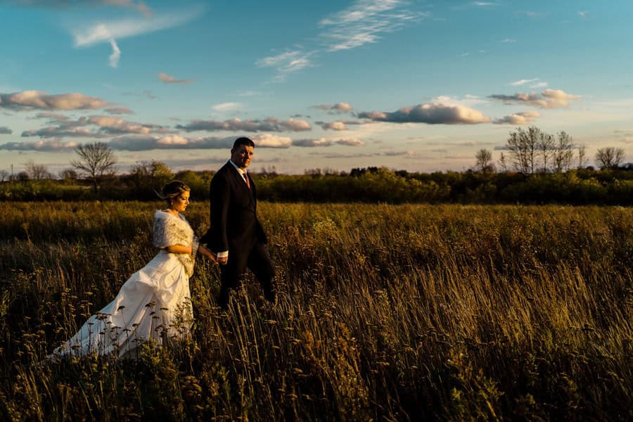 A bride and groom walking through a field at sunset.